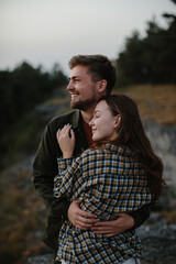 A handsome young man and a pretty woman in each other's arms are enjoying the sunset in the mountains.