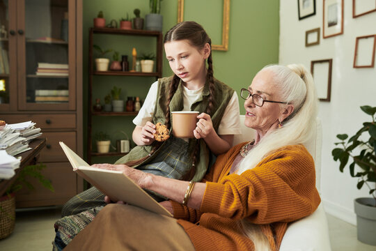 Elderly woman with long gray hair sharing stories from a book with young girl holding cookie and drink, surrounded by plants and books in a cozy room