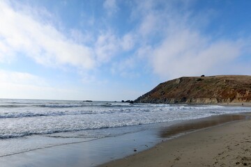 Pacifica Beach, California, with golden sands and gentle waves meeting the Pacific Ocean. A small mountain rises in the background, framed by clear skies, creating a peaceful coastal scene.