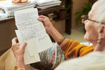 Naklejka premium Elderly person reading an intricately handwritten letter, sitting beside a cozy desk filled with stacked papers and plants, expressing emotions of nostalgia and longing while reminiscing