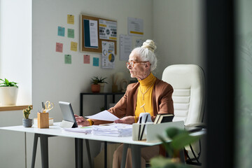 Elderly woman engaged in paperwork while seated at desk in a contemporary home office, with various documents and stationery spread out across workspace