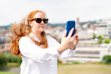 Young Woman With Flowing Red Hair Enjoying Sunny Day While Taking Selfie in City
