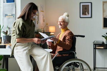 Collaborative work between elderly woman in wheelchair and young woman standing nearby. Brightly lit modern office with shelves, plants, and contemporary artwork in background