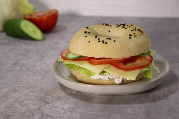 Delicious bagel with vegan cheese and fresh vegetables on a plate, grey table blurry background