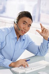 Young asian male customer service representative wearing headset working at desk in office, looking...