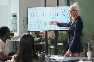 Senior businesswoman presenting data analysis on large screen to diverse team in modern office meeting room filled with charts and documents