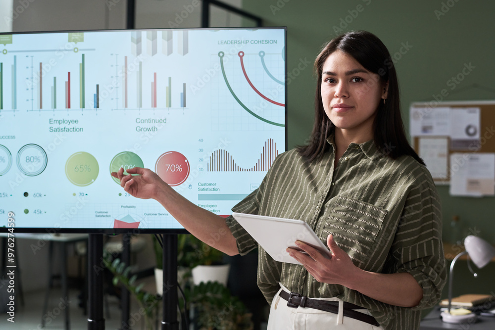 Poster Woman pointing at a presentation with business metrics displayed on screen, holding a tablet and showcasing various growth indicators in office setting