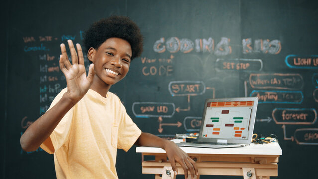 Smart african boy use laptop to code program and turn around to wave hand. Cute highschool stident coding engineering prompt or program system at blackboard in STEM technology classroom. Edification