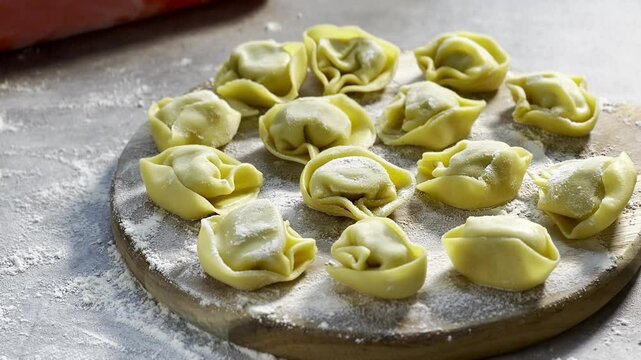 Raw traditional ravioli tortellini on wooden plate, grey table with flour