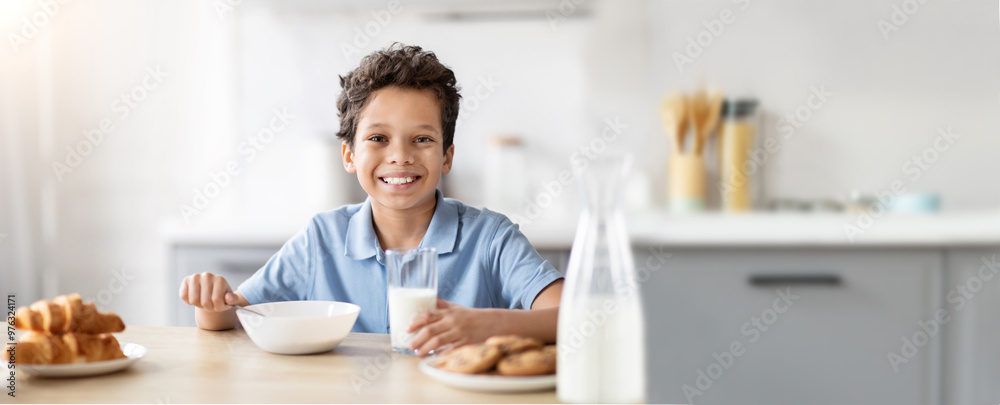 Wall mural a cheerful young african american boy sits at a kitchen table, smiling as he holds a glass of milk a