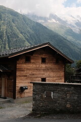 A small wooden house in a mountainous area. In the background, towering green mountains covered with forests and some fog are visible, creating a serene, alpine atmosphere.