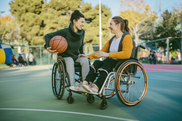 Obraz premium Two women in wheelchairs smiling and enjoying a basketball game on an outdoor court.