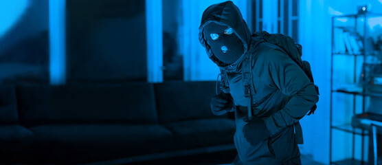 A figure dressed in dark clothing with a hood and mask stands in a dimly lit room at night. The atmosphere is tense, with blue lighting highlighting the scene.