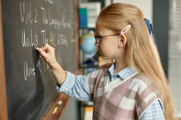 Side view of studious blonde schoolgirl wearing eyeglasses writing on blackboard during lesson at...