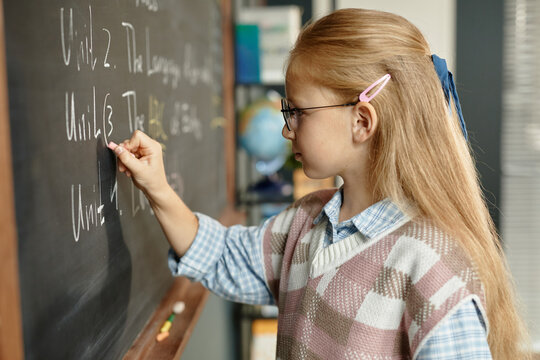 Side view of studious blonde schoolgirl wearing eyeglasses writing on blackboard during lesson at primary school, copy space