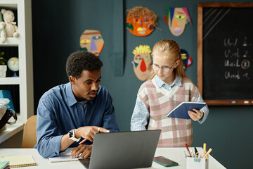 Medium shot of experienced male primary school teacher of African American ethnicity sitting at table talking to young smart student while pointing at laptop screen in classroom, copy space