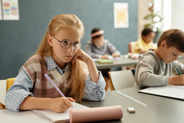 Medium shot of studious young girl with glasses sitting at desk attentively looking at blackboard...