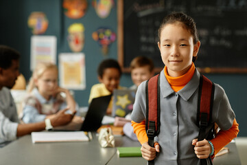 Portrait of young Central Asian schoolgirl with adorable smile wearing casual clothes and backpack while posing in primary school classroom, copy space