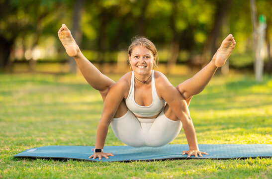 Smiling sporty fit young girl performing Tittibhasana or Firefly pose, arm-balancing asana with legs stretched out forwards during morning yoga practice in green summer park