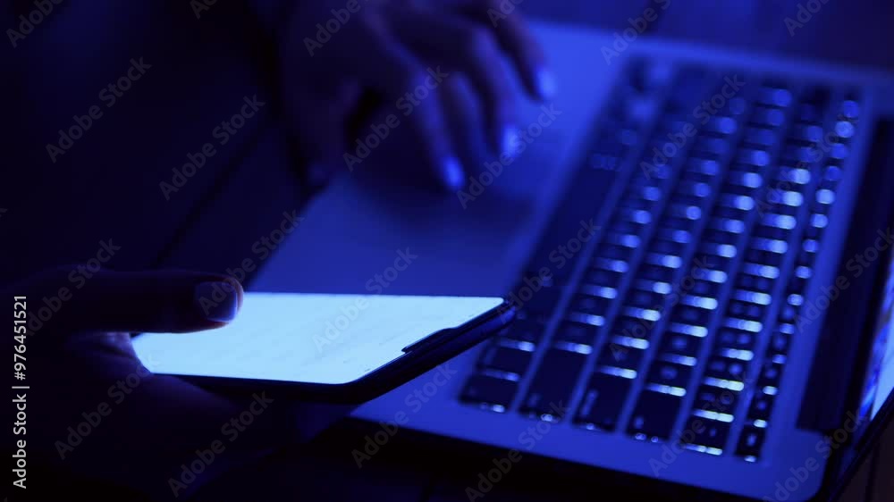 Poster A person types on a laptop with one hand while holding a smartphone in the other, illuminated by a faint blue light.