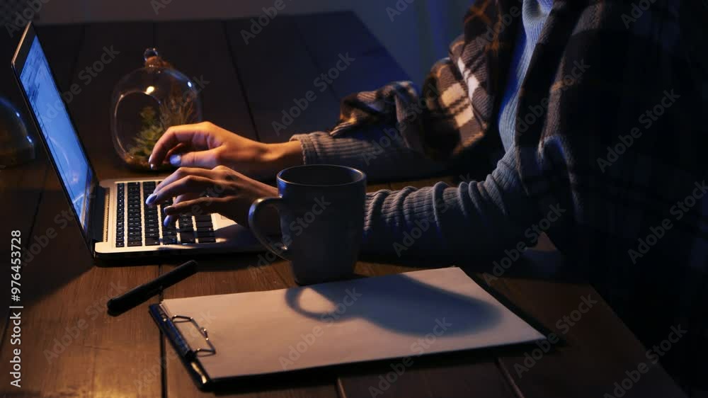 Poster A person works on a laptop in low light, with a mug and notepad nearby, creating a cozy atmosphere.