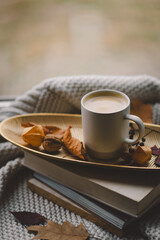 A warm cup of coffee sits atop a golden tray adorned with dried leaves, resting on a stack of books, with a cozy blanket draping nearby.