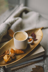 A warm cup of coffee sits atop a golden tray adorned with dried leaves, resting on a stack of books, with a cozy blanket draping nearby.
