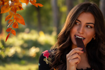 Fototapeta premium Closeup shot of amazing beautiful brunette biting dark chocolate in the park, fall season 