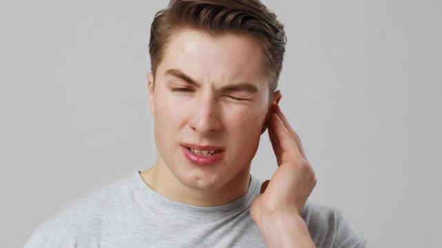 Otitis. Studio portrait of young man suffering from ear pain, close up, grey background