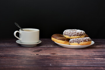 A cup of hot coffee and a plate with several fresh donuts in chocolate and caramel glaze on a dark wooden table.