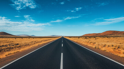 Naklejka premium A long, straight highway stretches through a vast, arid desert under a clear blue sky, flanked by dry grass and distant hills.