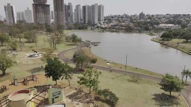 Drone flies over lake in park toward large buildings in Campo Grande, Mato Grosso do Sul, Brazil