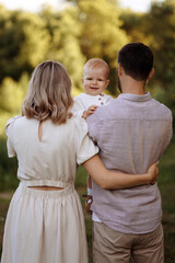 A family of three, a man and a woman with a child in their arms, stand in a park in the summer. The concept of a happy family with a small child, care and love.