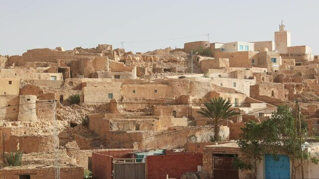 Traditional Berber earthen houses with characteristic flat roofs, nestled on hillside of Tamezret village under clear sky with minaret of hilltop mosque in background, Tunisia