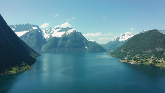 Aerial view of a deep blue fjord surrounded by towering cliffs and mountains in Norway, under a bright sky with coastal community on edge