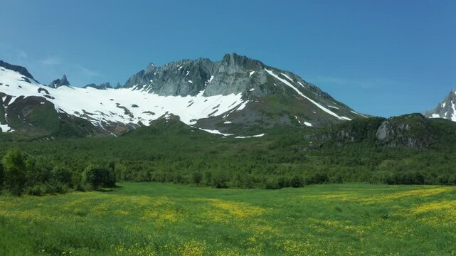 Aerial dolly of a snow-capped mountain in Norway, surrounded by lush green fields and a clear blue sky during summer
