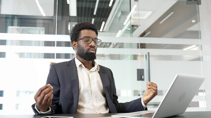 African American businessman in a formal suit practicing meditation with closed eyes in office to reduce stress. Professional in suit engaging in mindfulness at workplace. Calm worker practices yoga