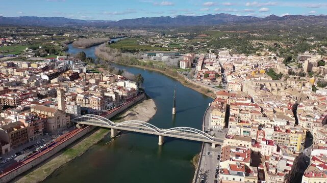 Aerial view of Catalan city of Tortosa on sunny spring day overlooking reconstructed Bridge of State across Ebro river and Monument to Battle of Ebro in middle of water, Spain