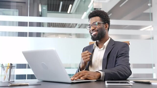 Happy handsome african american businessman in formal suit receiving great news on laptop sitting at desk in business office. Satisfied smiling entrepreneur celebrating victory showing yes gesture