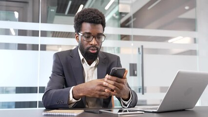 Upset african american businessman in formal suit receiving bad news on phone while sitting at a desk at a workplace in a business office. Concerned black man reads an unpleasant message on smartphone - Powered by Adobe