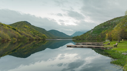 Obraz premium A photo of a serene outdoor scene with a calm lake reflecting the sky and surrounding green mountains. The sky is overcast, with a few clouds. The mountains have a forest of trees growing on them.