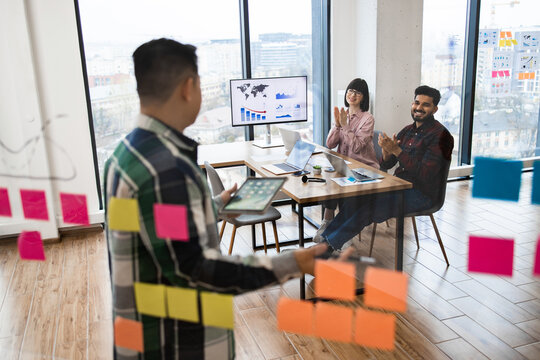 Business team meeting in modern office with view. Colleagues interacting, brainstorming with tablet and sticky notes. Graphs, charts on screen. Collaborative environment with natural light.