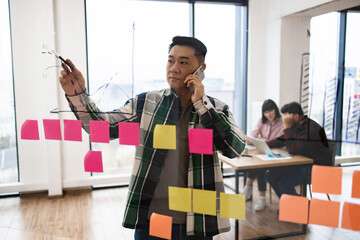 Business professional using phone and writing on glass board with colorful sticky notes. Planning, brainstorming, and strategizing in modern office.
