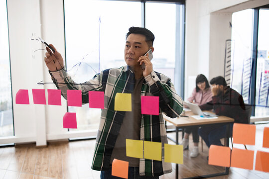 Business professional using phone and writing on glass board with colorful sticky notes. Planning, brainstorming, and strategizing in modern office.