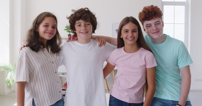 Four smiling Caucasian classmates or friends in casual clothes standing together in classroom and hugging each other. Cheerful high school children looking at camera, demonstrating close friendship.