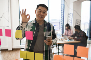Smiling man in casual office setting giving okay hand sign while holding smartphone. Team collaboration and brainstorming session with sticky notes on glass wall.