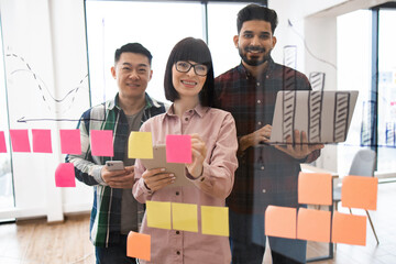 Diverse team collaborating during brainstorming session. Three people using sticky notes, charts, smartphones, and laptops in office setting. Concept of teamwork, creativity, and innovation.