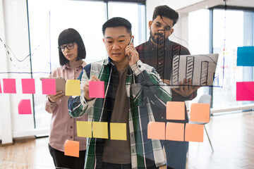 Team members brainstorming creative ideas on transparent board using sticky notes and electronic devices. Professional collaboration and planning in modern office environment