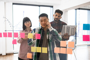 Group of diverse colleagues brainstorming ideas using sticky notes and chart drawings on glass wall. Creative team planning new project together in modern office.