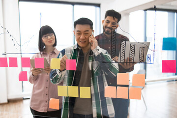 Multiracial team collaborating during business meeting with colorful sticky notes and bar charts. Coworkers brainstorming strategy ideas on transparent board, each person in project planning.